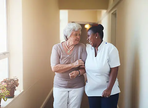 AdobeStock_112047591 - Blue Water Homecare Caregiver laughs with elderly woman while escorting her down a hall